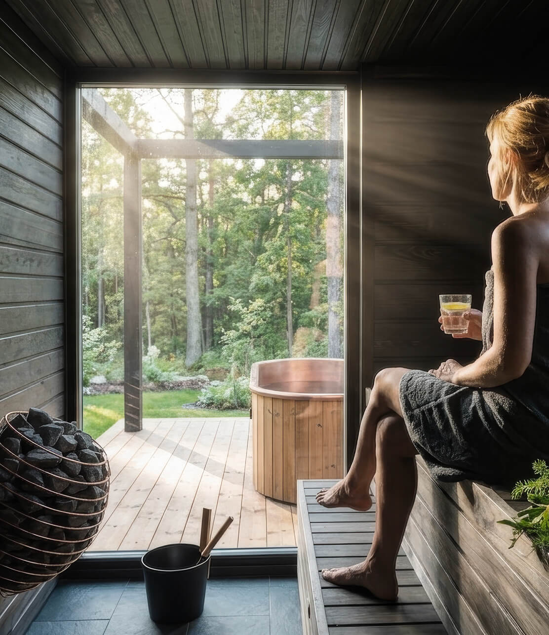 A woman seated in a dark timber Löyly&Stone sauna looking through a full-height window onto a green garden